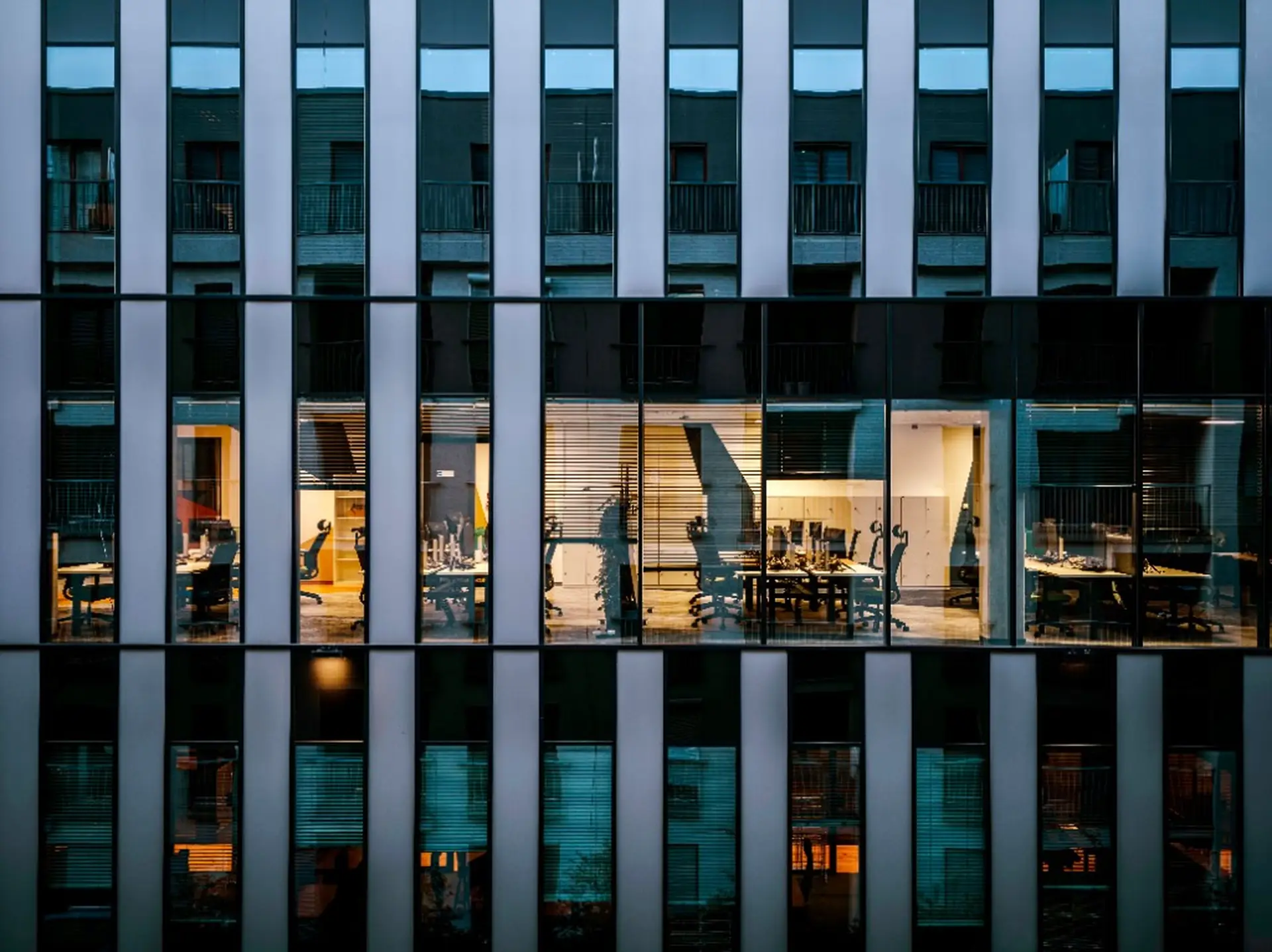 Twilight office building facade with a lit floor of desks and monitors seen through the windows