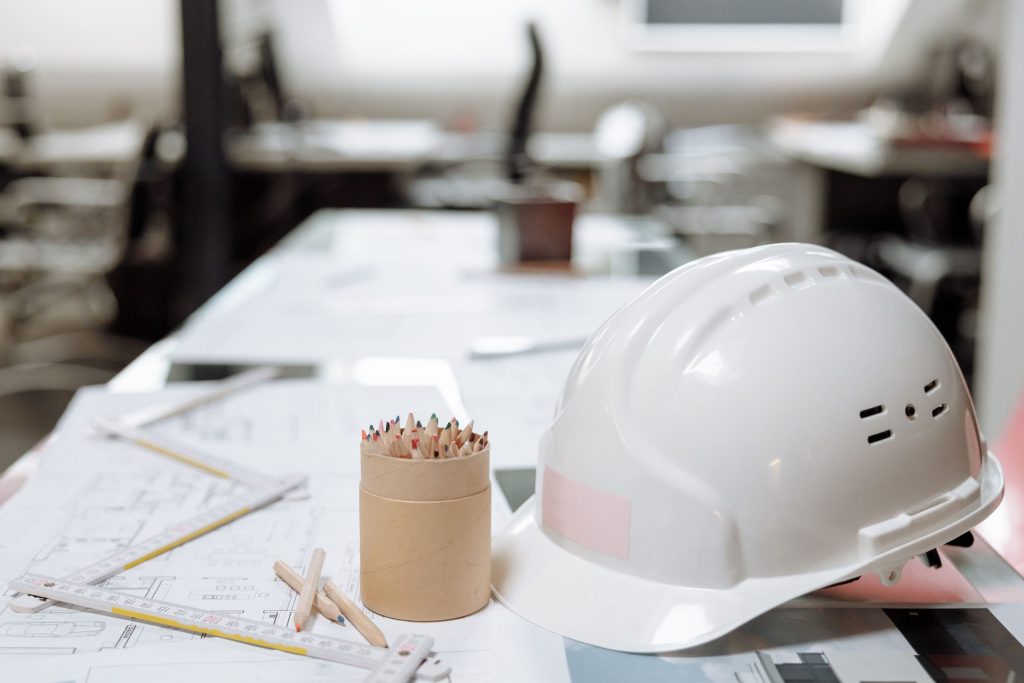 White construction safety helmet placed on architectural blueprints with drafting tools and colored pencils on an engineer's office desk.