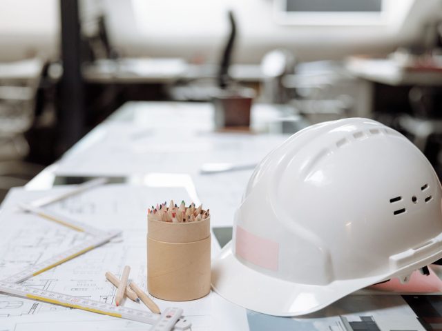 White construction safety helmet placed on architectural blueprints with drafting tools and colored pencils on an engineer's office desk.