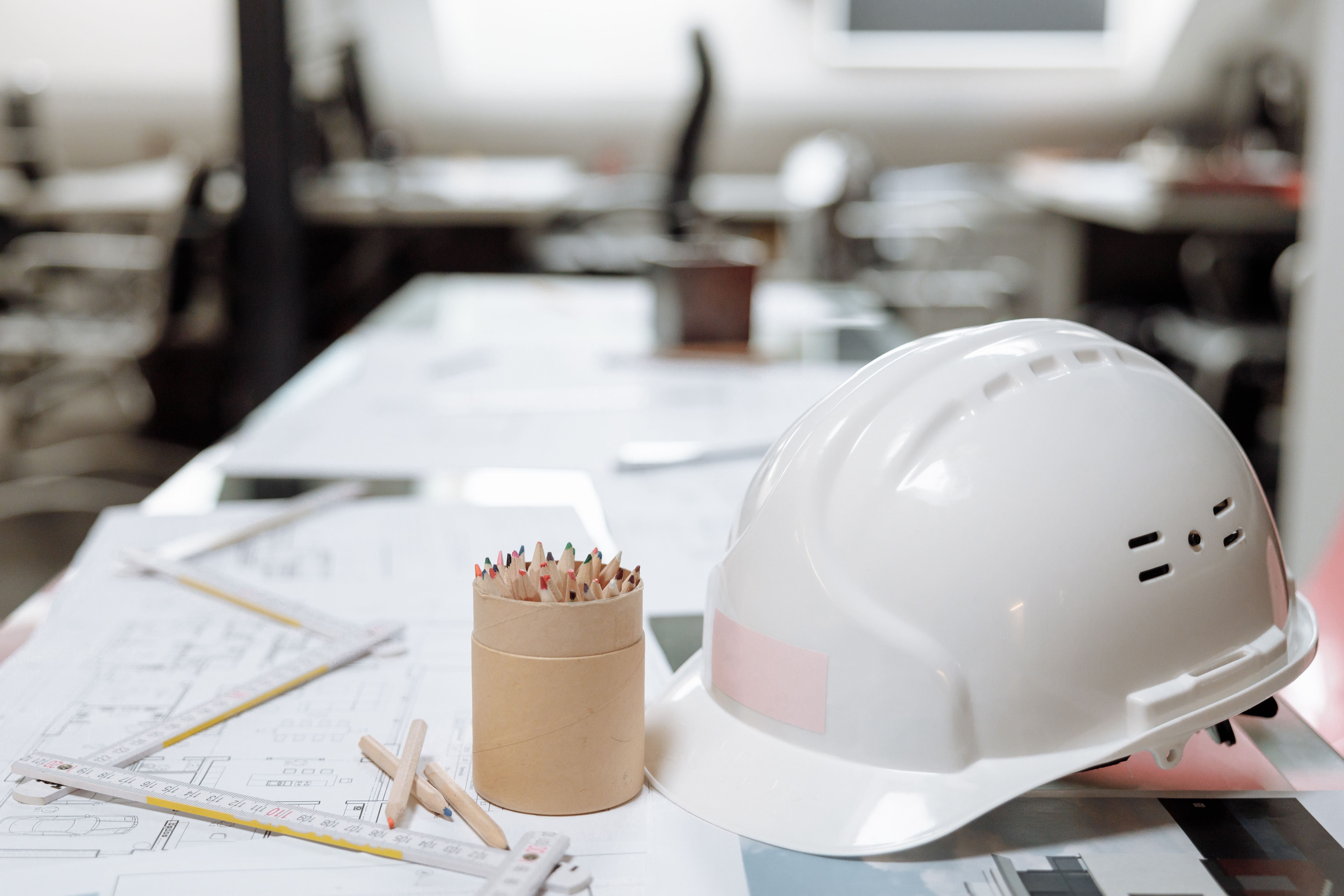 White construction safety helmet placed on architectural blueprints with drafting tools and colored pencils on an engineer's office desk.