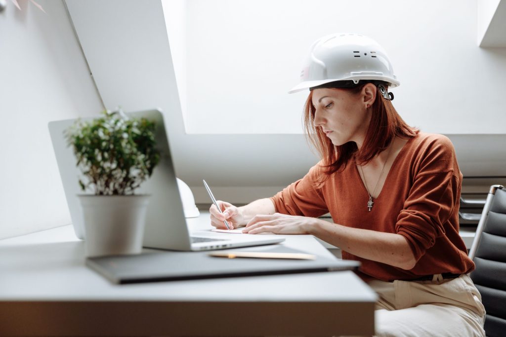 Construction engineer reviewing project documents and taking notes while working on a laptop in a modern office, wearing a safety hard hat.