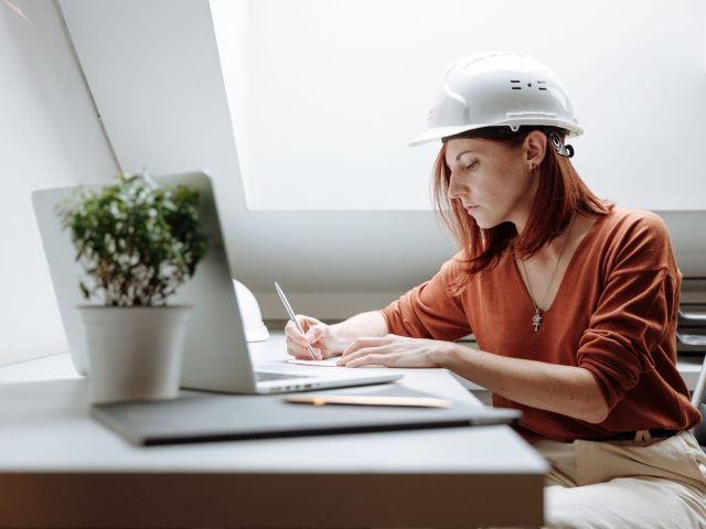 Construction engineer reviewing project documents and taking notes while working on a laptop in a modern office, wearing a safety hard hat.