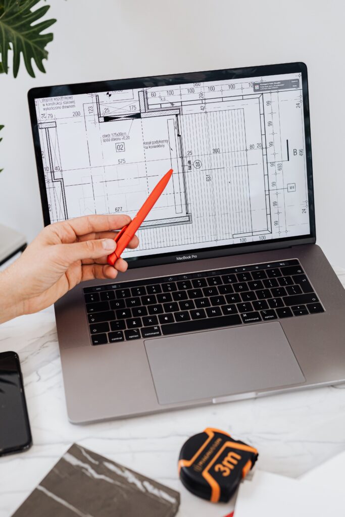 CAD drafter reviewing a detailed architectural floor plan on a laptop, pointing to construction dimensions and layout specifications during a design review.