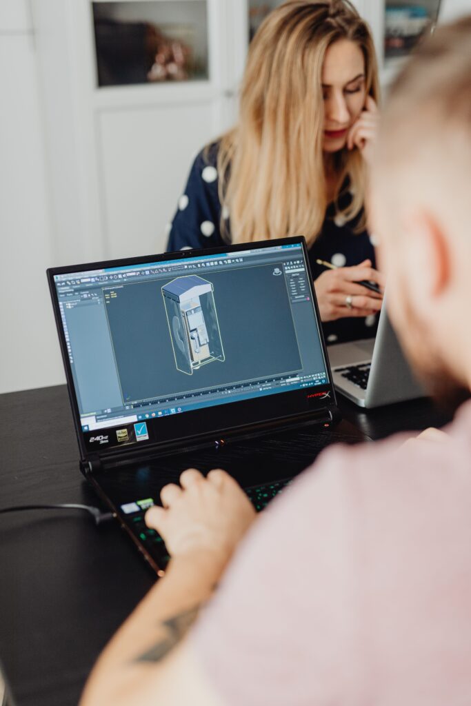 Engineer using a laptop to work on a 3D CAD model while collaborating with a colleague in an office environment.