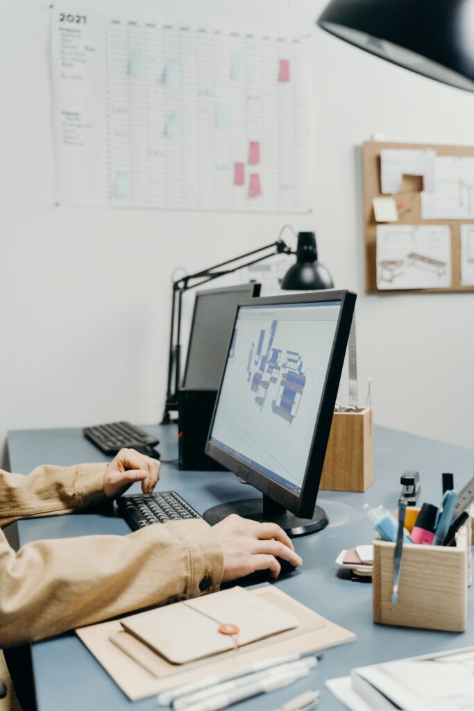 Person working at a desk using CAD software on a desktop computer, reviewing a 3D design model in a modern engineering workspace with project planning charts in the background.