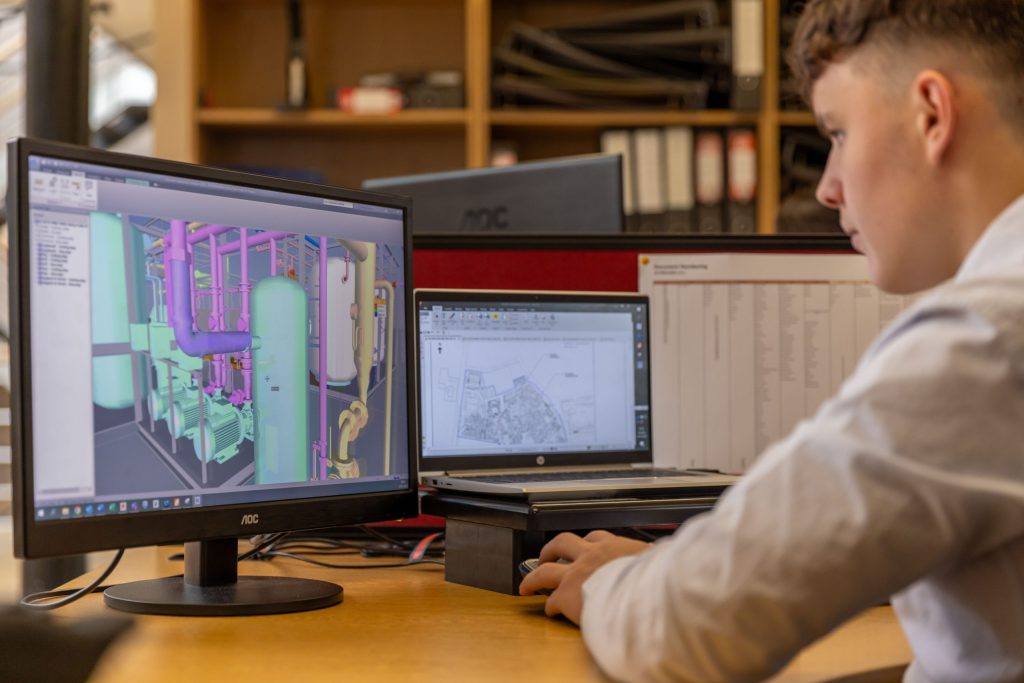 CAD technician reviewing a detailed BIM model of mechanical piping systems on a desktop monitor in a professional engineering office.