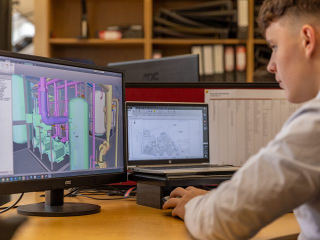 CAD technician reviewing a detailed BIM model of mechanical piping systems on a desktop monitor in a professional engineering office.