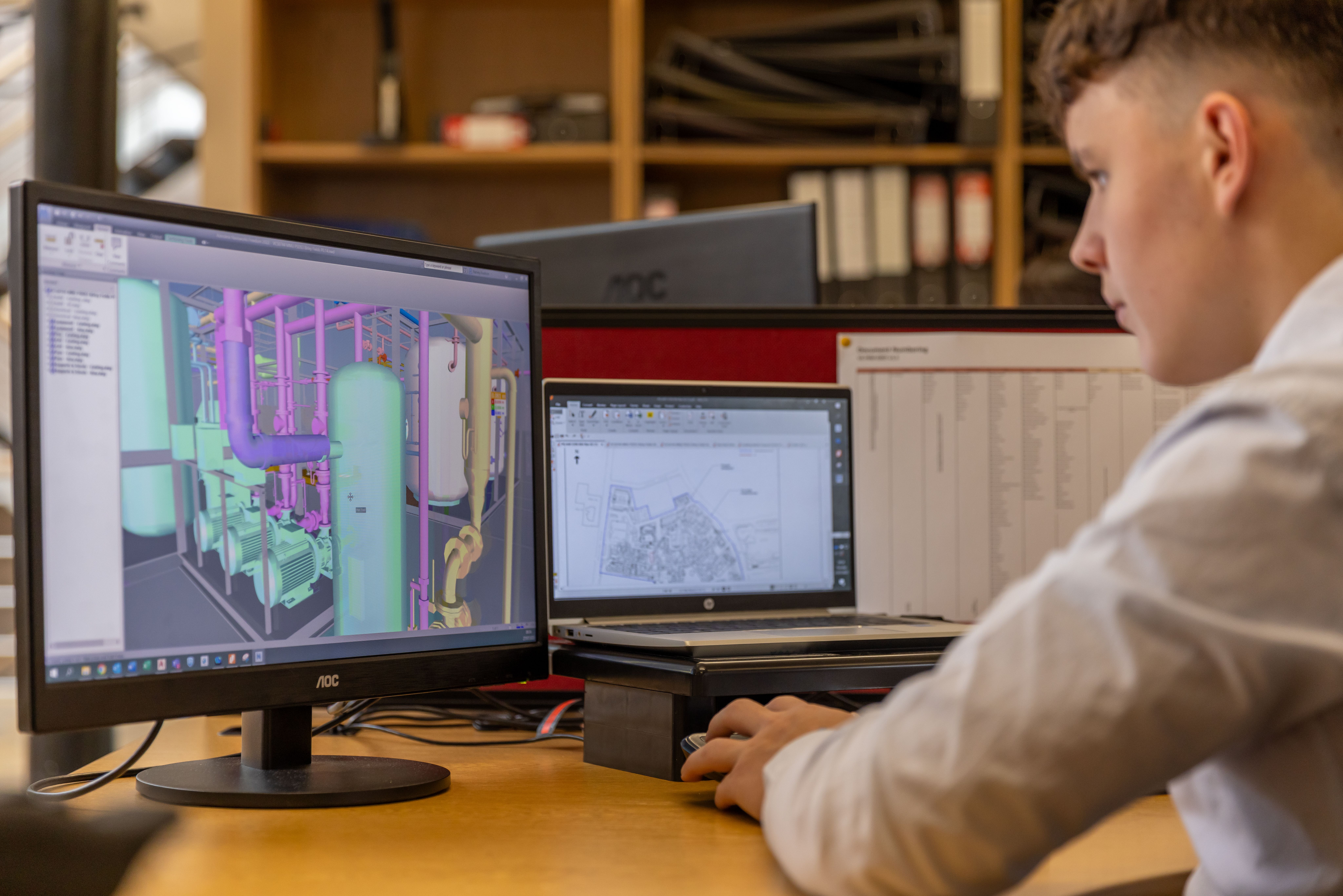 CAD technician reviewing a detailed BIM model of mechanical piping systems on a desktop monitor in a professional engineering office.