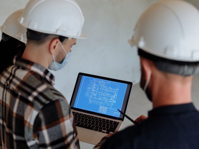 Construction engineers reviewing a digital blueprint on a laptop at a project site while wearing safety helmets.