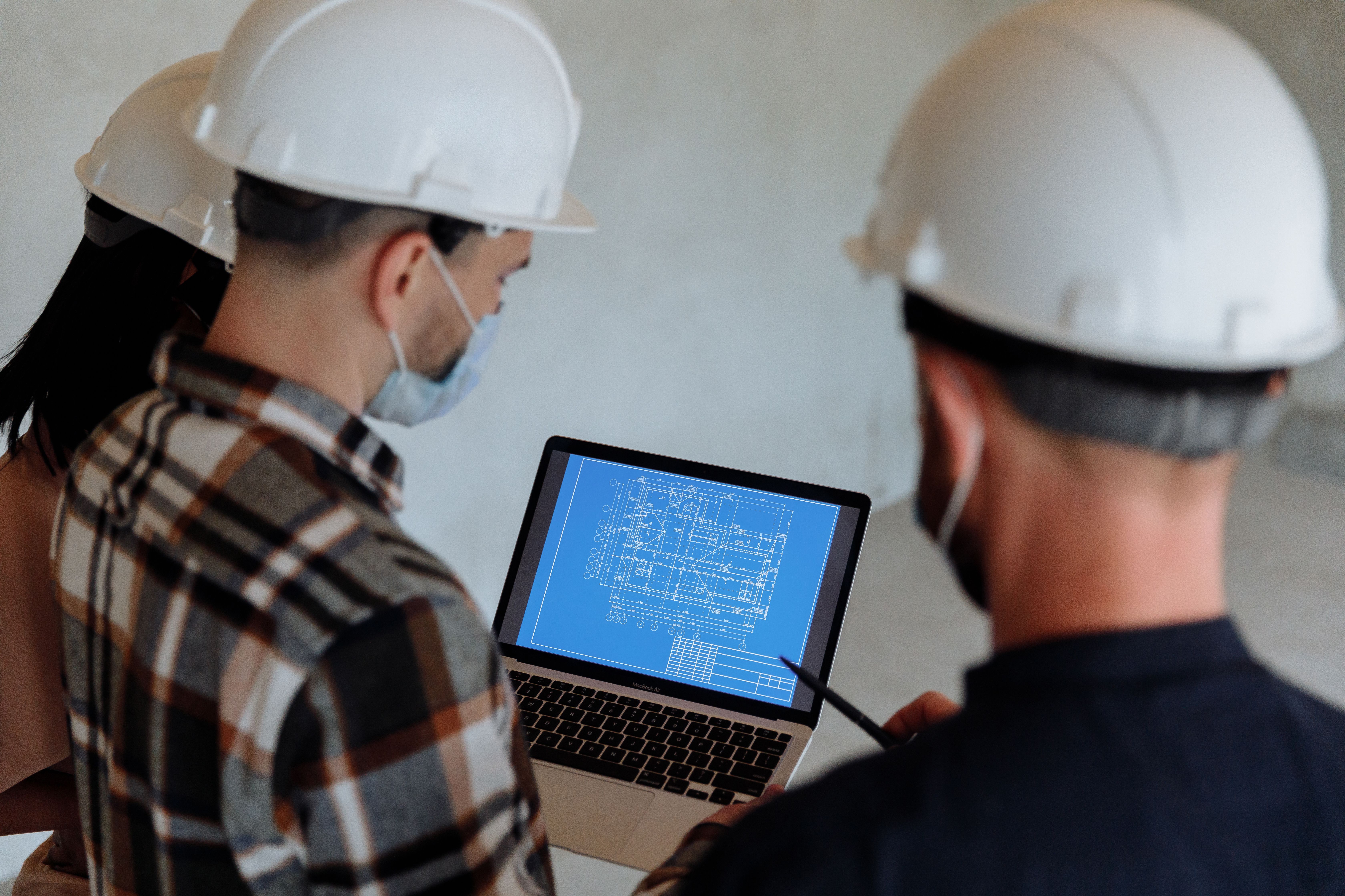 Construction engineers reviewing a digital blueprint on a laptop at a project site while wearing safety helmets.