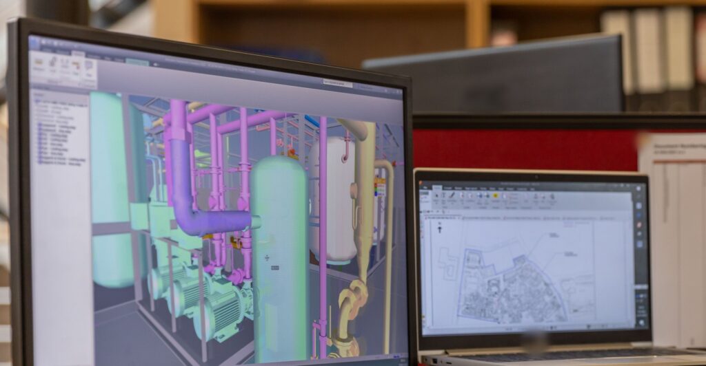 BIM specialist workstation showing a coordinated 3D MEP pipework model next to a site plan on a laptop