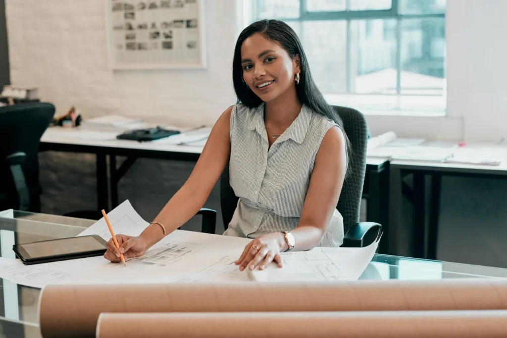 Virtual assistant for architects marking up floor plan drawings at an architecture studio desk, with a tablet and rolled blueprints nearby