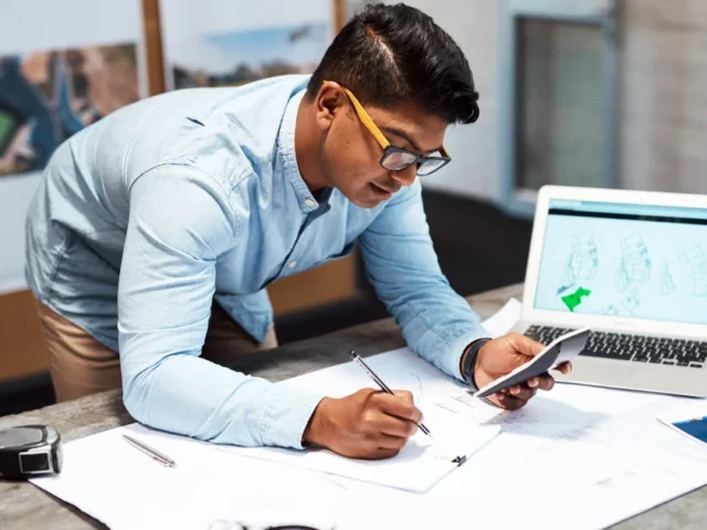 South Asian construction estimator in a blue shirt reviewing blueprints with a laptop open on his office desk.