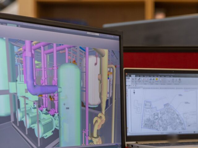 BIM specialist workstation showing a coordinated 3D MEP pipework model next to a site plan on a laptop