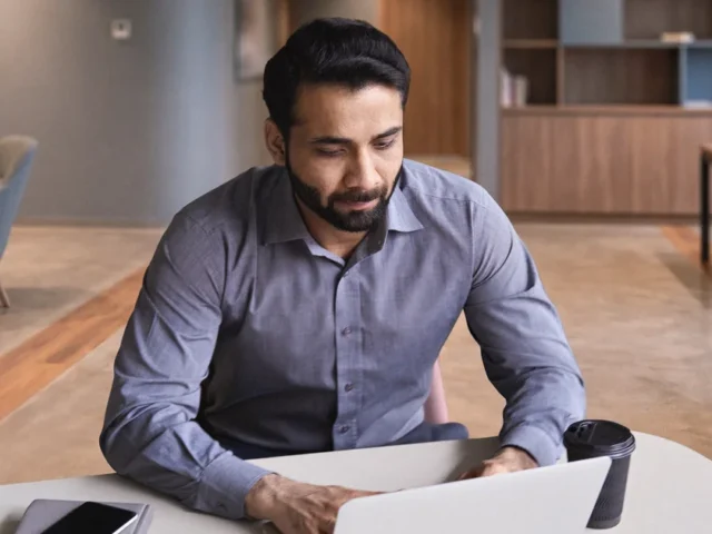 South Asian virtual assistant in a business shirt working on a laptop at a desk in a modern office.