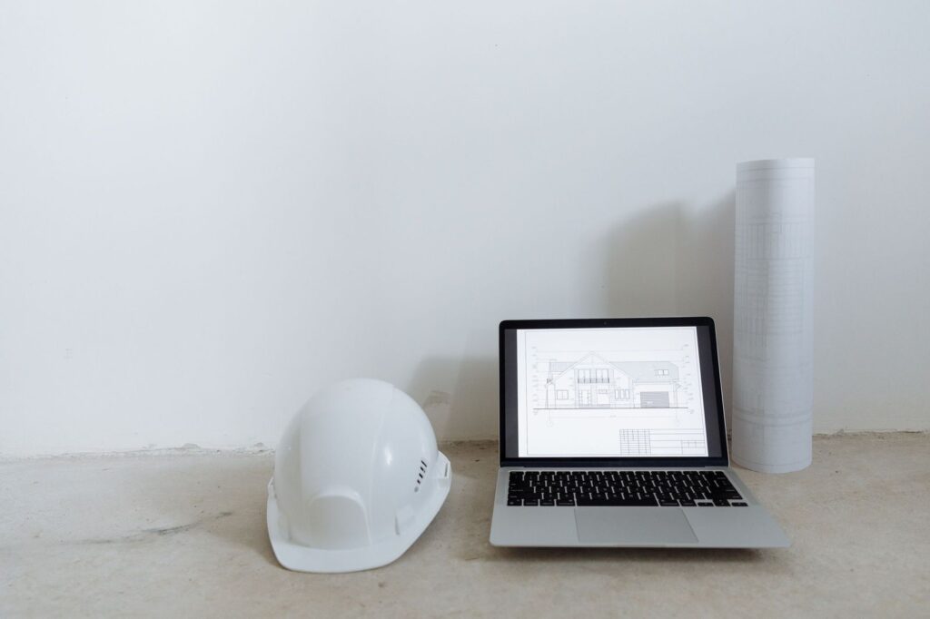 Laptop showing construction drawings beside rolled plans and a hard hat, a shared setup for remote estimating